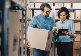 A man and woman are standing amongst shelving. The man holds a box and they are consulting a list.