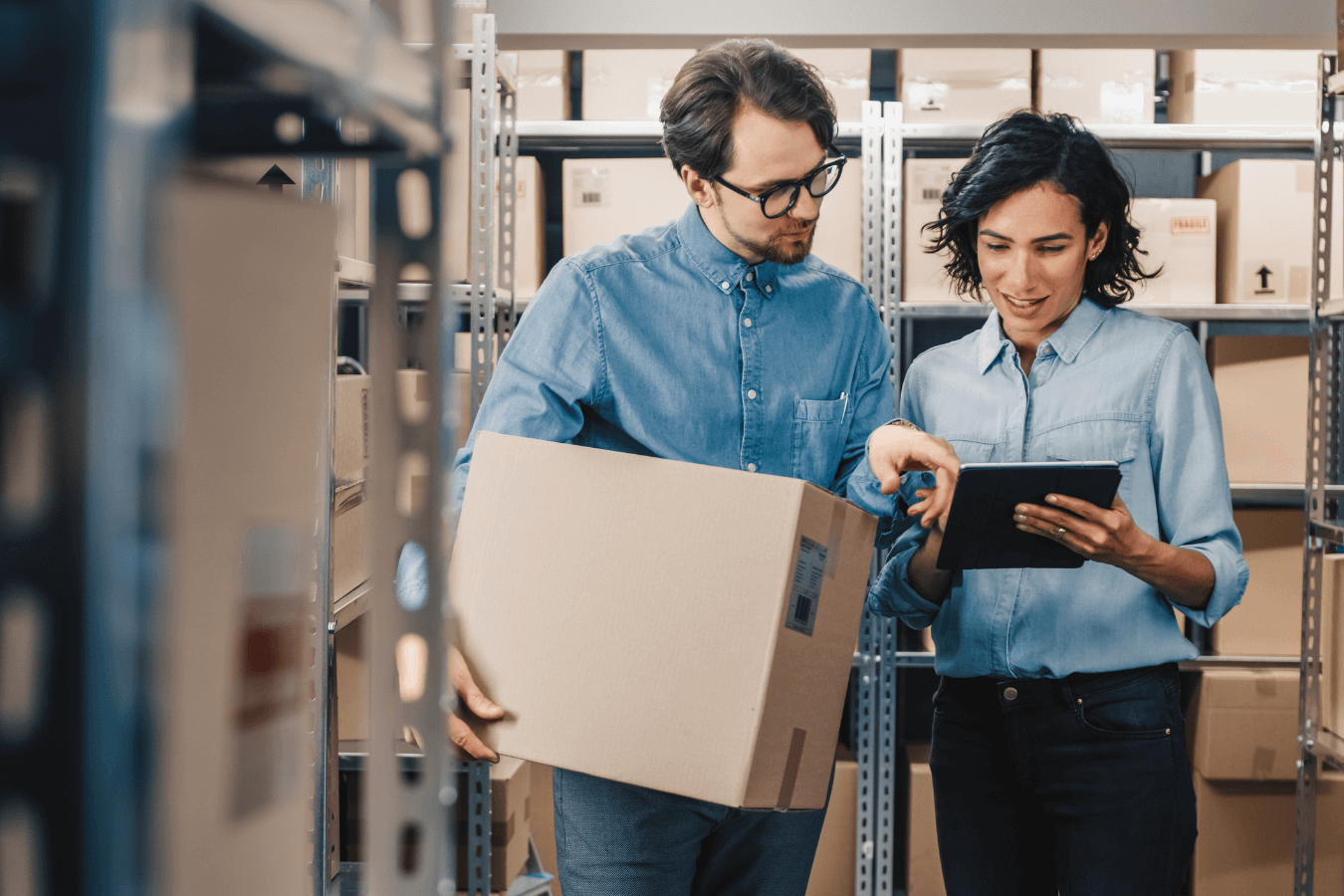 A man and woman are standing amongst shelving. The man holds a box and they are consulting a list.