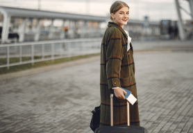 a professional woman wearing a tweed overcoat pulls a suitcase while looking over her shoulder at the camera.
