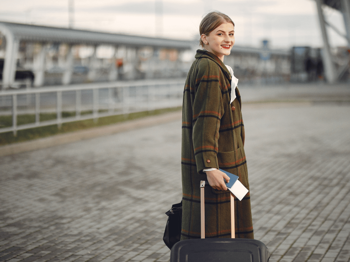 a professional woman wearing a tweed overcoat pulls a suitcase while looking over her shoulder at the camera.