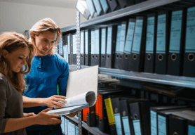 Two people look through a file in front of shelving containing lever arch files