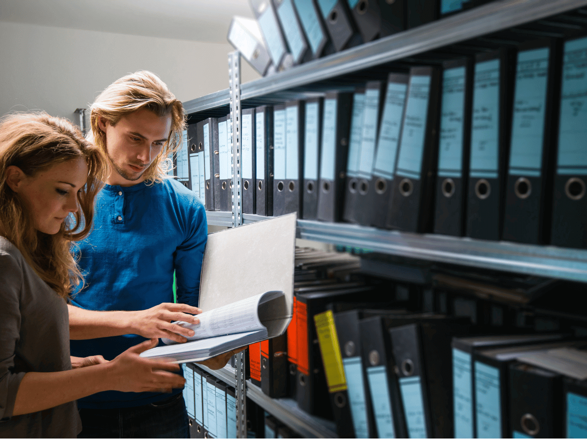 Two people look through a file in front of shelving containing lever arch files