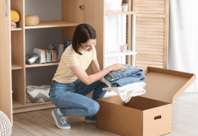 a woman in t-shirt and jeans packs away jeans and jumpers into a cardboard box ready for storage.