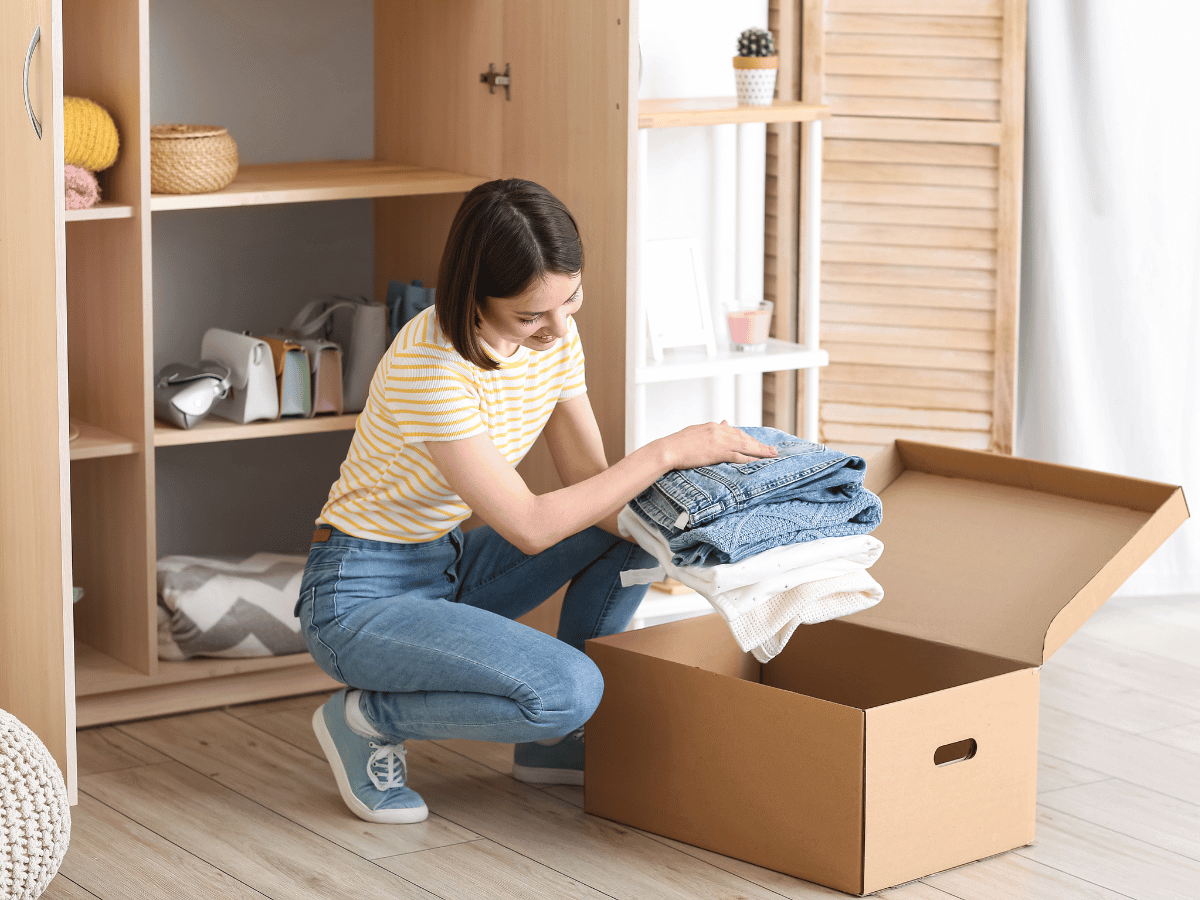 a woman in t-shirt and jeans packs away jeans and jumpers into a cardboard box ready for storage.