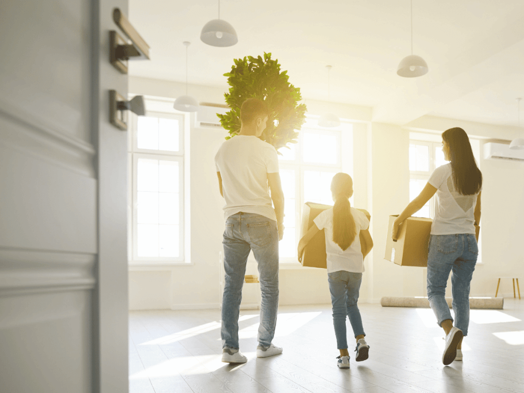 a family of three carry packed removal boxes after a quick home sale. The room is empty and sunlight is shining from the windows.