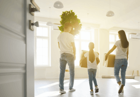 a family of three carry packed removal boxes after a quick home sale. The room is empty and sunlight is shining from the windows.